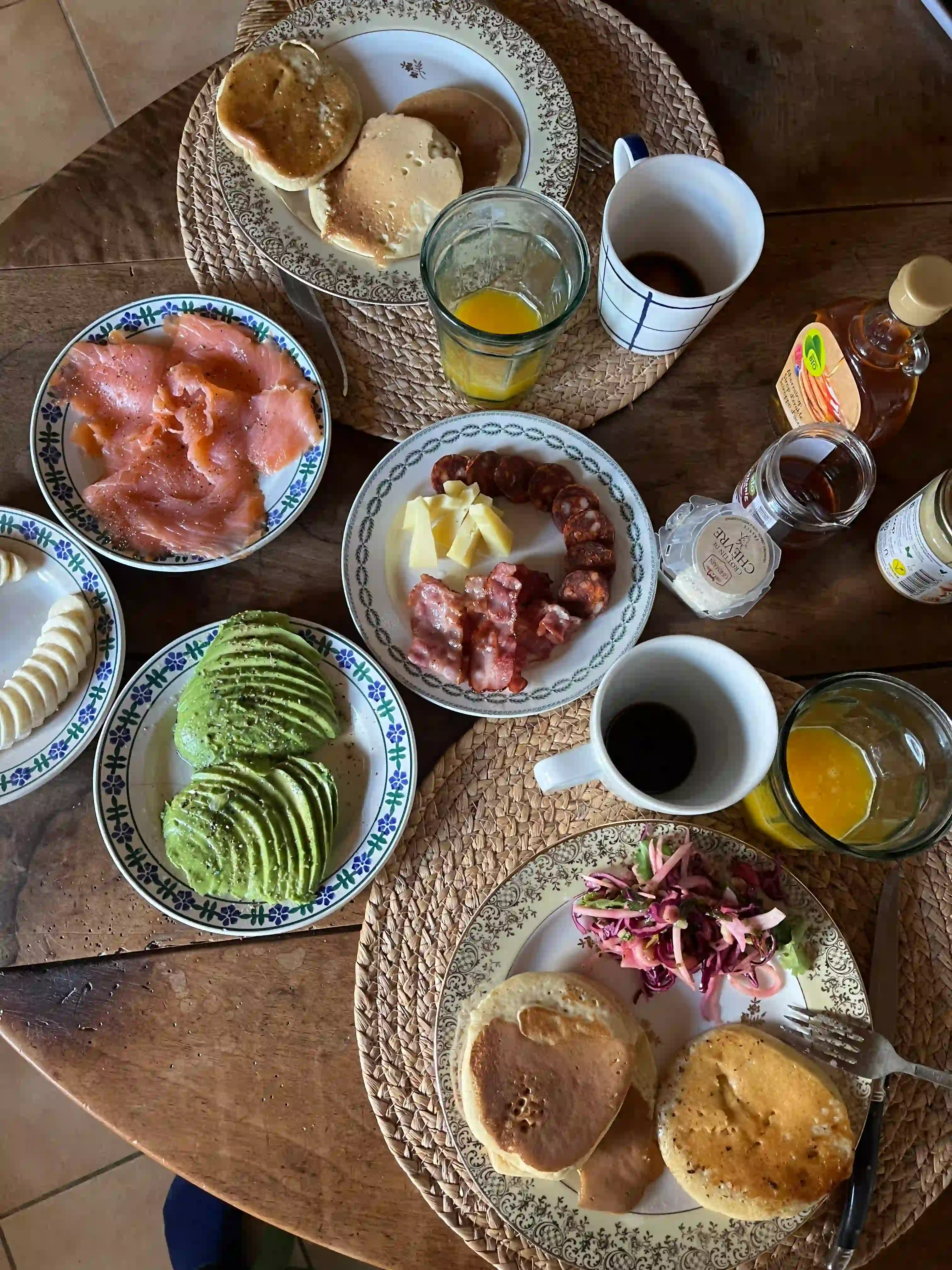 Breakfast table with pancakes, avocado slices, banana slices, smoked salmon, bacon with cheese, orange juice, coffee mugs, and condiments.