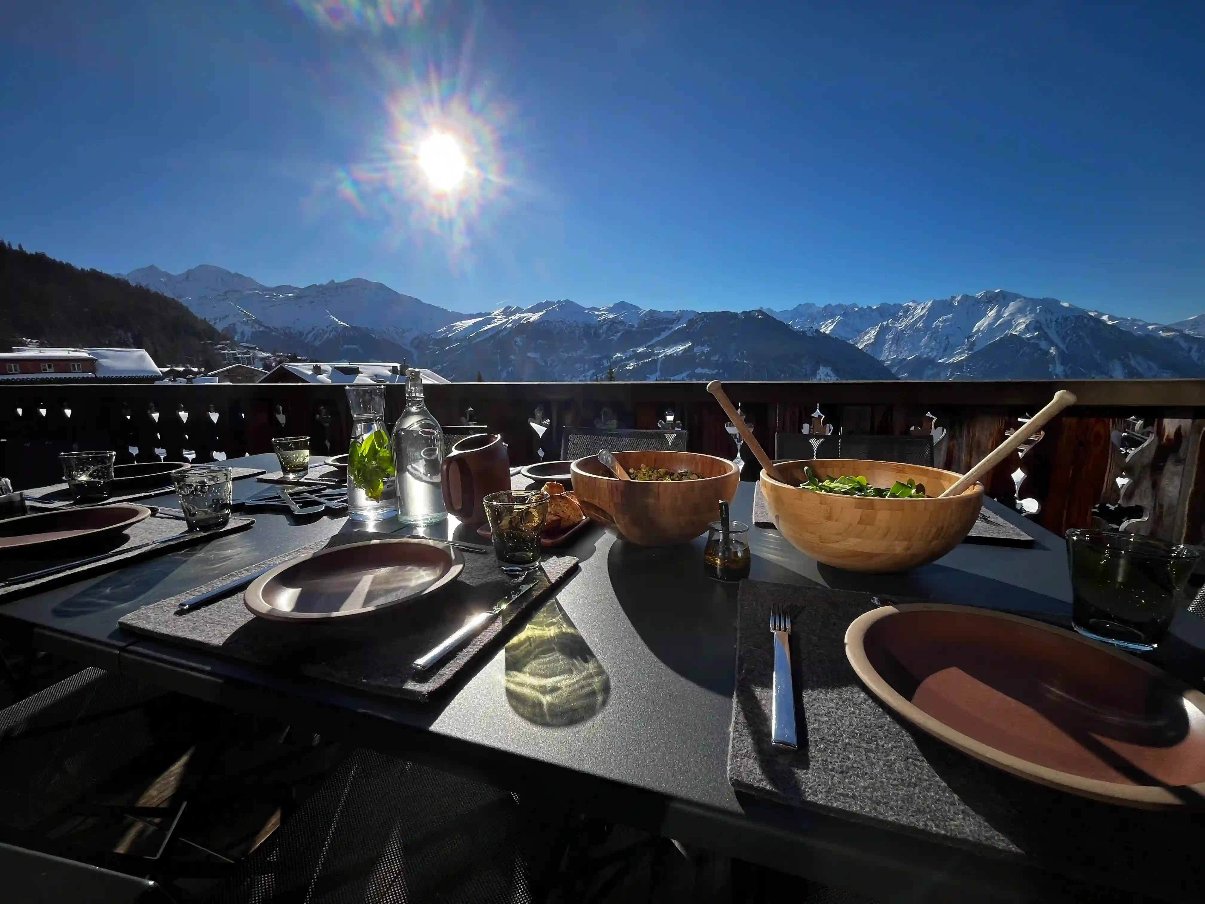 Outdoor dining table set with plates, wooden bowls of salad, and glasses under bright sun with snow-covered mountains in the background.