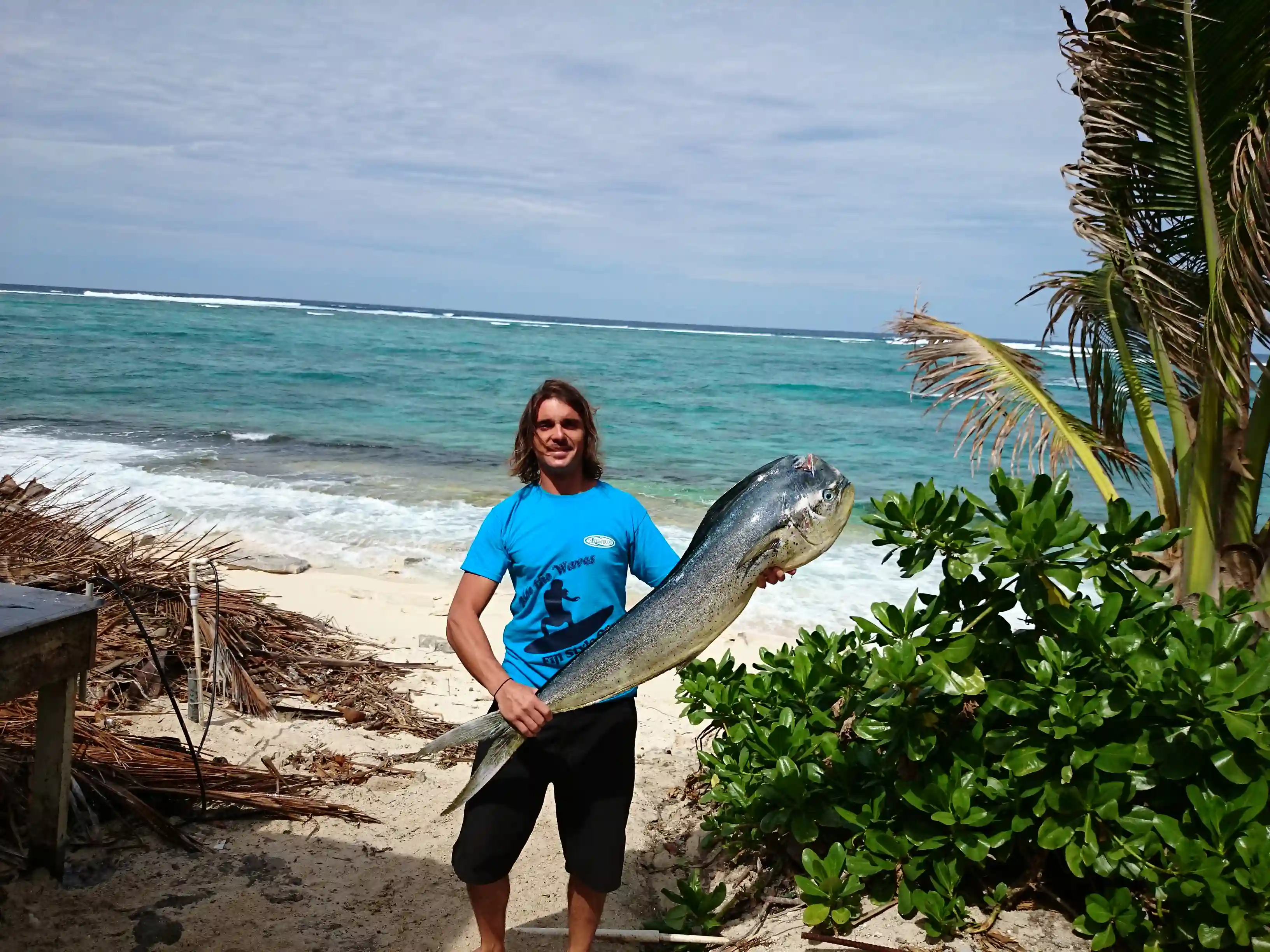 Man standing on beach holding a large dolphinfish with ocean waves and palm trees in the background.