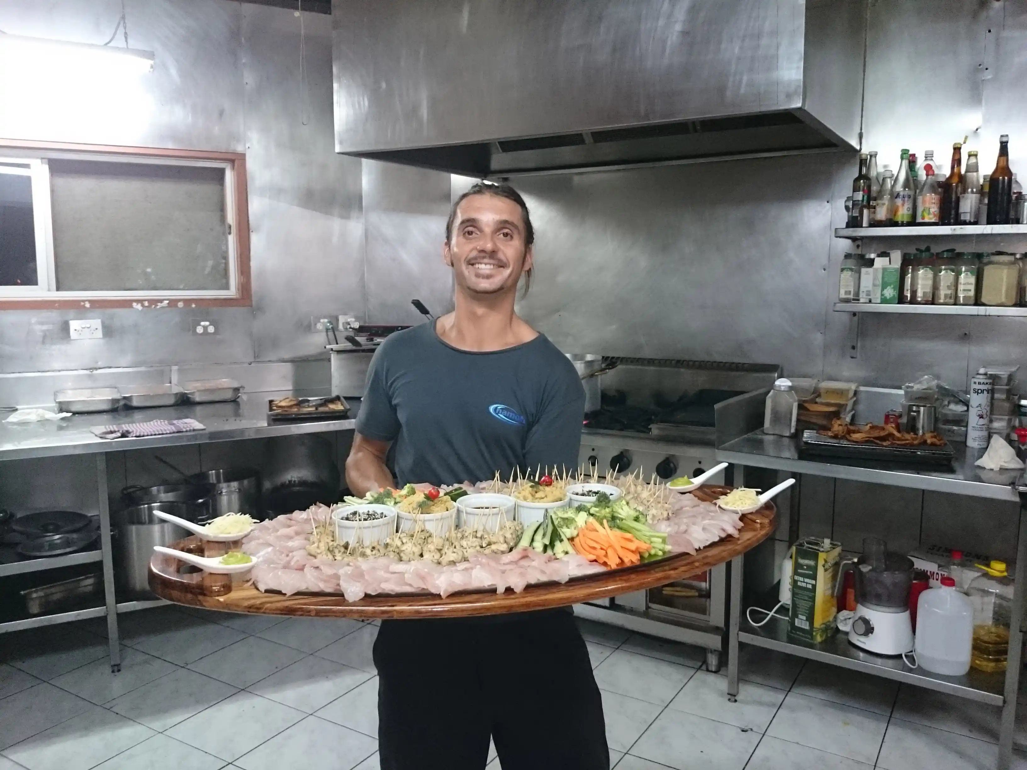 Smiling man holding a large wooden platter with an assortment of sliced fish, vegetables, and dipping sauces in a commercial kitchen.